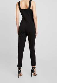 Kvinna som bär en ärmlös svart jumpsuit med dragkedja i ryggen och svarta högklackade skor med ankelrem, stående mot en enkel bakgrund.