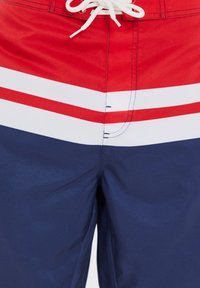 Pantaloni da bagno rossi e blu navy con strisce bianche; dotati di vita regolabile con cordoncino e tessuto leggero e liscio. Cuciture a macchina visibili.