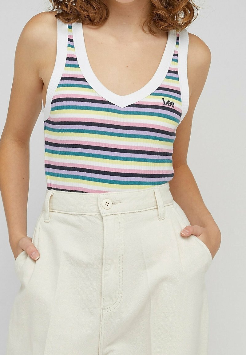 Femme portant un haut sans manches rayé multicolore à col en V avec un bord blanc et un pantalon crème taille haute, mains dans les poches.