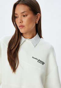 Felpa bianca con una cucitura diagonale, adornata con una stampa nera che dice "Sunday Running Club." Indossata sopra una camicia a righe con colletto.