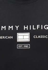 Fekete anyag fehér felirattal, amelyen az áll: "Tommy Hilfiger American Classic" egy téglalap alakú logó körül, alatta pedig "EST. 1985".