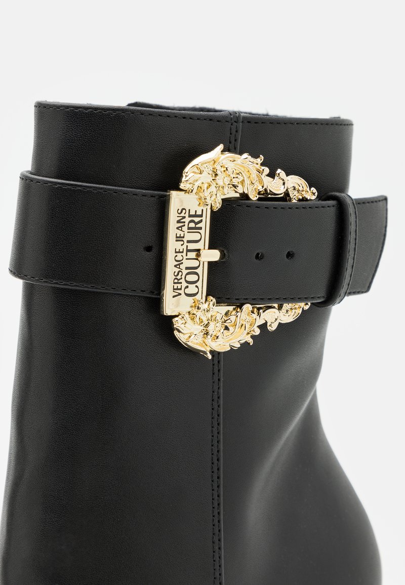 Stivale alla caviglia in pelle nera con una fascia larga e una fibbia decorativa dorata, caratterizzato da dettagli intricati. Texture liscia con una forma elegante.