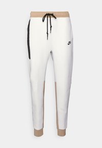 Hvide Nike joggingbukser med beige linning og manchetter, sort snøre og sort sidelomme med lynlås på venstre ben.