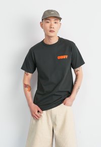 Black cotton t-shirt with an orange "OBEY" logo on the left chest, paired with beige plaid shorts. Simple design and relaxed fit.
