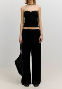Zwarte strapless top van textuurstof, gecombineerd met losse zwarte broek. Model houdt een zwarte schoudertas met een gladde afwerking.