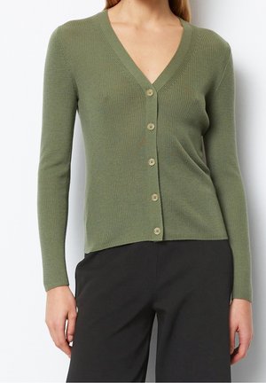 Frau trägt eine grüne, gerippte Strickjacke mit Knöpfen, V-Ausschnitt und langen Ärmeln, kombiniert mit schwarzen Hosen vor einem einfarbigen Hintergrund.