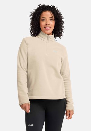 Vrouw met krullend haar, gekleed in een beige fleece met kwart-ritssluiting en zwarte broek, met een ontspannen uitdrukking op een lichte achtergrond.