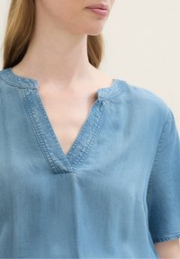 Vrouw in een lichtblauwe spijkerblouse met korte mouwen en een V-vormige gestikte halslijn, afgebeeld van de nek tot halverwege de torso tegen een neutrale achtergrond.