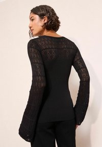 Juoda ilga rankovė viršus, turintis lygiagretų kūną ir sudėtingas nėriniuotas raštuotas rankoves su flaro detale. Neutralus fonas.