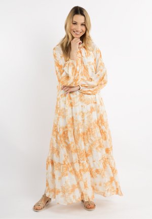 Lächelnde Frau steht mit der Hand nahe am Kinn, trägt ein langes weiß-orangen gemustertes Kleid und beige Sandalen vor einfarbigem Hintergrund.