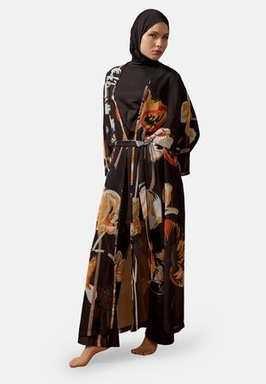 Kvinde iført sort hijab og en lang sort blomsterkjole med orange, beige og hvide blomster, stående barfodet mod en ensfarvet baggrund.