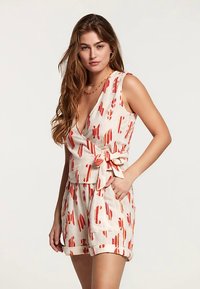 Viegls krēmkrāsas linu kombinezons ar sarkaniem abstraktiem rakstiem. Raksturīgas V veida apkakle, regulējama jostas lente un uzruļļoti piedurkņu gali ērtai pieguvei.