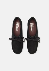 Juodi suede tipo mokasinai su apvalia nosimi, raištelių dekoracija ir kontrastingomis rudos spalvos vidpadžiais, ant kurių įspausta "Clarks ORIGINALS".