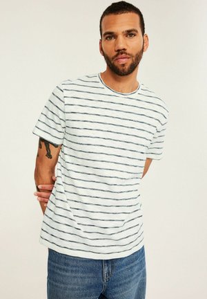 Baard dragende man die een wit T-shirt met dunne zwarte horizontale strepen en een blauwe spijkerbroek draagt, staat met de handen achter zijn rug.