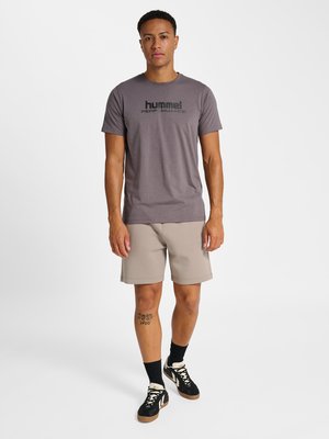 Mand iført grå "hummel performance" T-shirt, beige shorts, sorte sokker og sorte sneakers, stående mod en ensfarvet hvid baggrund.