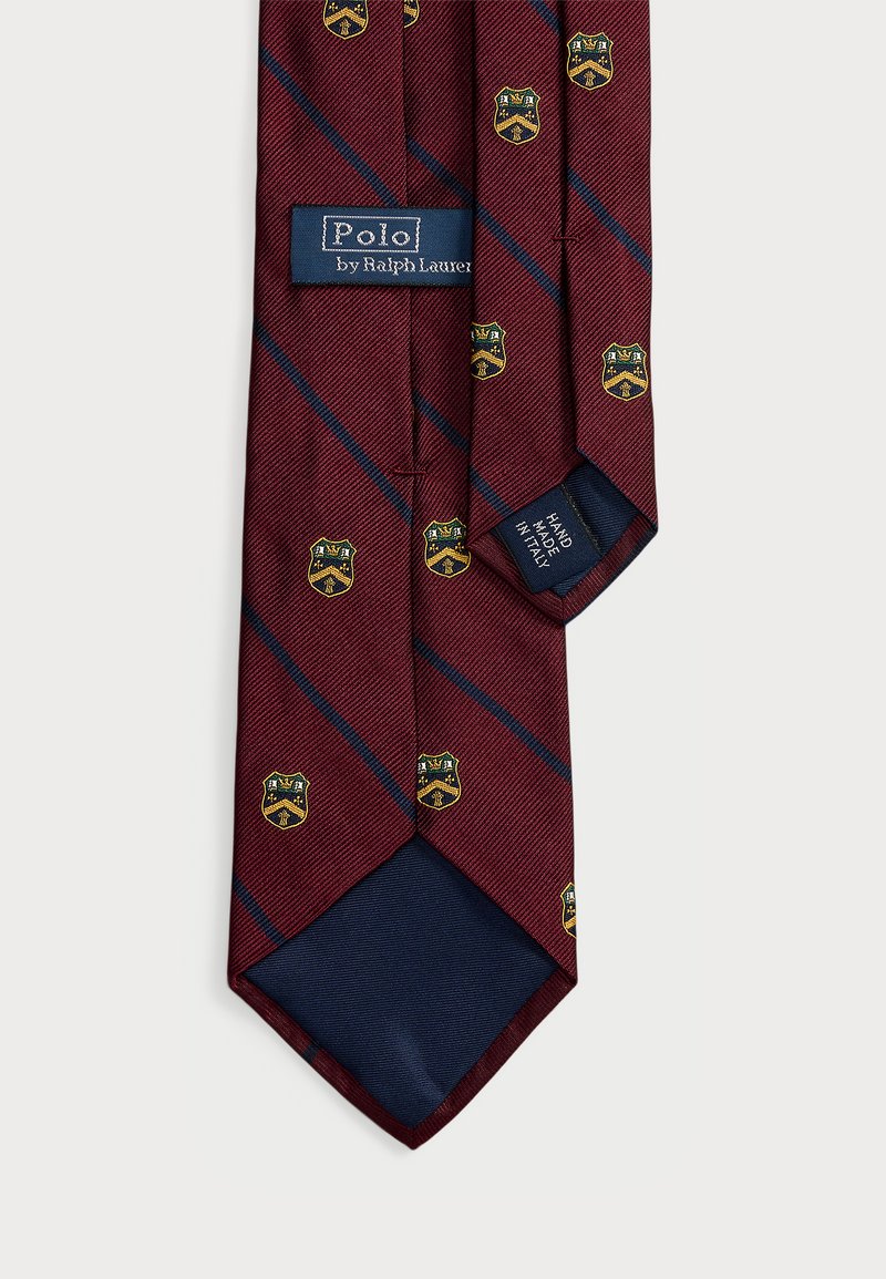 Corbata burdeos con rayas diagonales en azul marino, adornada con motivos de escudos en dorado y azul. Hecha de una tela texturizada con etiqueta hecha a mano.