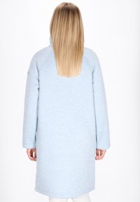 Donna con lunghi capelli biondi che indossa un cappotto blu chiaro fino al ginocchio, di spalle su uno sfondo bianco uniforme.