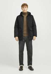 Chaqueta negra con capucha y dos bolsillos frontales; combinada con un suéter marrón de punto trenzado y jeans de mezclilla oscuros. Los zapatos son oscuros y con cordones.