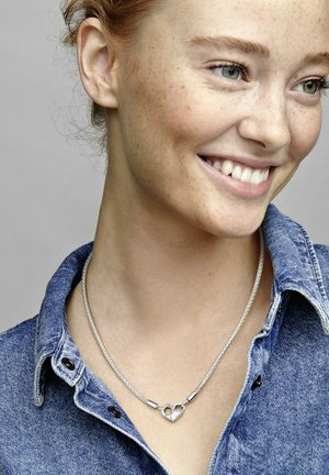 Silbernes geflochtenes Halsband mit herzförmigem Verschluss, getragen über einem Jeanshemd. Besitzt eine glatte metallische Textur und einen dezenten Glanz.