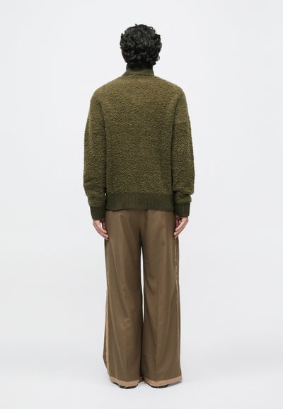 Olivegrüner, strukturierten Pullover mit hohem Kragen, kombiniert mit weiten braunen Hosen, die über eine Seitenstreifen-Detail verfügen. Vom Rücken betrachtet.