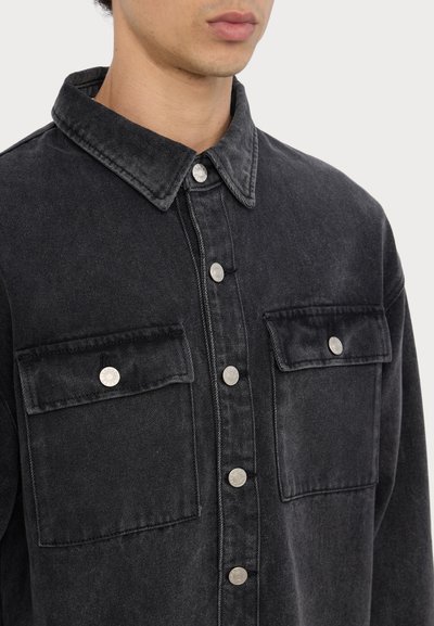 Zwarte spijkerblouse met een kraag, zilveren drukknoopsluitingen, twee borstzakken en een zachte, gestructureerde afwerking.