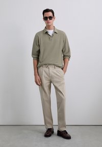 Polo de manga larga verde claro, pantalones beiges y zapatos marrones oscuros. Diseño oversize con cuello abotonado y tejido texturizado.