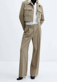 Veste beige courte avec deux poches avant, chemise blanche en dessous et pantalon beige à jambes larges ; des chaussures noires complètent l'ensemble.