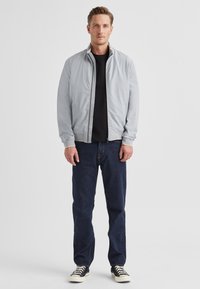 Graue Bomberjacke mit Stehkragen, durchgehendem Reißverschluss, gerippten Bündchen, getragen über einem schwarzen Hemd, kombiniert mit dunkelblauen Jeans und schwarzen Turnschuhen.