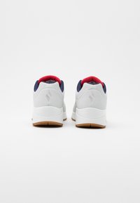 White athletic shoes with navy accents and red lining. Features a sleek design, textured upper, and a rubber outsole with grooves.