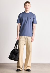 Μπλε βαμβακερό μπλουζάκι με στρογγυλή λαιμόκοψη και μικρό λογότυπο στο στήθος, συνδυασμένο με μπεζ τζιν παντελόνι φαρδιάς γραμμής και μαύρη τσάντα.