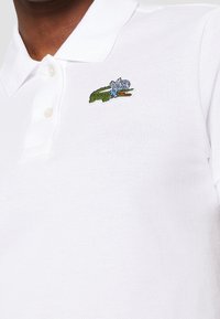 Valkoinen polo-paita, joka on valmistettu teksturoidusta kankaasta. Vasemmassa rintataskussa on vihreä ja sininen brodeerattu krokotiililogot. Kauluksessa on kolme nappia.