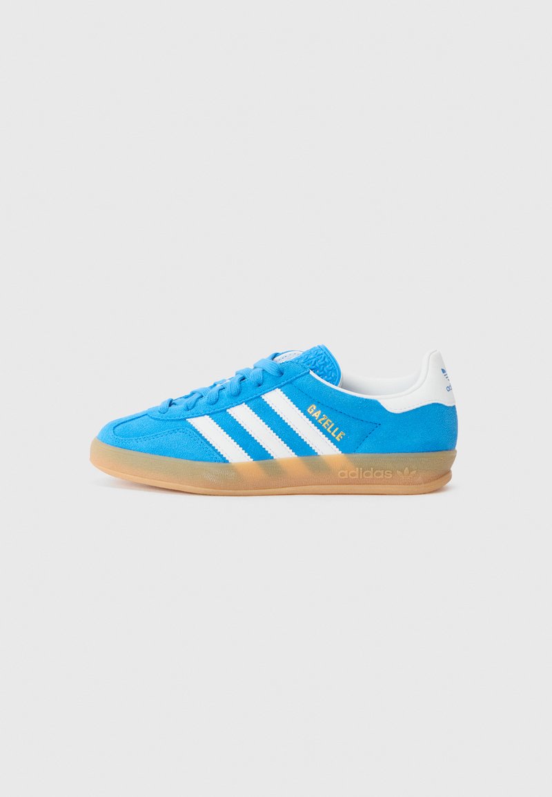 Sapatilhas de camurça azul com riscas brancas, língua texturizada e sola de borracha de goma. Apresenta o logótipo "Gazelle" em dourado na lateral.