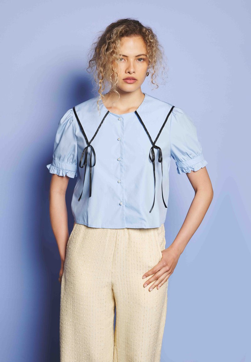 Ung kvinne med krøllete hår iført lyseblå bluse med puffermer og svarte bånddetaljer sammen med kremfargede teksturerte vide bukser.