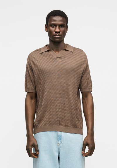 Homme portant un polo en maille texturée marron à manches courtes et un pantalon en denim bleu clair, debout devant un fond blanc uni.