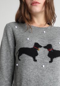 Femme portant un pull gris avec deux silhouettes de chien noires se faisant face, décorées de pompons blancs et de petits accents rouges et dorés.