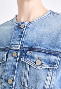 Chaqueta de mezclilla azul claro con escote redondo, cierre frontal con botones, dos bolsillos en el pecho y detalles en herrajes de tono plateado.