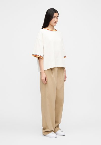 Krémovo sfarbené nadrozmerné tričko s červeným a oranžovým lemom, v kombinácii s voľnými hnedými nohavicami a bielymi teniskami. Mäkká látka, ležérny strih.