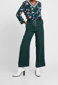 Blus med blommönster och lång ärm i grönt med V-ringning, ihop med vida byxor i mörkgrönt och vita högklackade ankelstövlar.