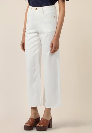 Kobieta w białych, szerokich spodniach z wysokim stanem i surowym wykończeniem nogawek oraz brązowych platformowych sandałach na obcasie, stojąca na beżowym tle.