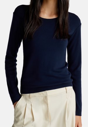 Vrouw draagt een nauwsluitende marineblauwe trui met lange mouwen, ingestopt in hooggetailleerde, crème kleurige, geplooide broek, met handen in de zakken.