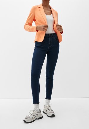 Person wearing a peach blazer, white tank top, dark skinny jeans, white socks, and chunky white sneakers standing against a plain background.