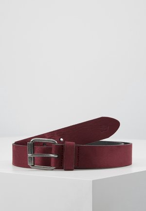 Ceinture en cuir rouge foncé avec boucle en métal argenté, enroulée sur une surface blanche sur un fond gris clair uni.