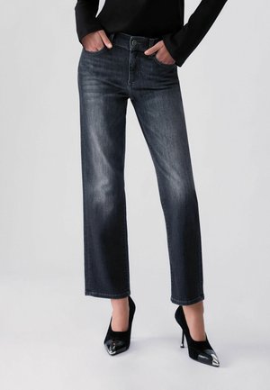 Osoba nosząca ciemne jeansy o prostych nogawkach, z rękami w kieszeniach i czarne buty na wysokim obcasie z metalowymi, ostro zakończonymi noskami.