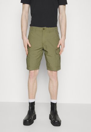 Pantaloni cargo verdi realizzati in un tessuto dalla trama liscia, con tasche laterali e chiusura a bottone. Abbinati a stivali neri alla caviglia e calzini bianchi.