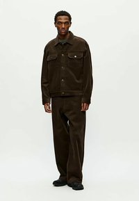 Modèle masculin portant une veste en velours côtelé marron foncé et un pantalon large assorti, se tenant contre un fond clair.
