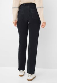 Jean droit noir à taille mi-haute, avec deux poches arrière et une texture lisse. Porté avec des baskets blanches et argentées.