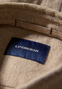 Interiör i beige tyg med synliga sömmdetaljer. Blå tygetikett med texten "LINDENBERGH" i vita bokstäver. Mjuk och jämn textur överlag.