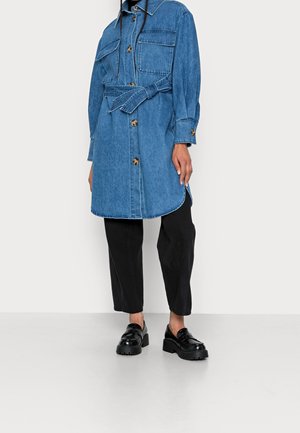 Vrouw die een blauwe denimjas met riem en voorzakken draagt, zwarte cropped broek en zwarte chunky loafers, staand tegen een witte achtergrond.