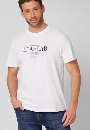 Uomo con indosso una maglietta bianca con la scritta "LEAF LAB", abbinata a jeans blu, in piedi davanti a uno sfondo semplice grigio chiaro.