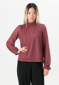Jonge vrouw met steil blond haar, gekleed in een langmouwige, gemêleerde bordeauxrode blouse en zwarte broek, poseert met één hand op haar heup.